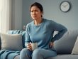 Person sitting on a couch at home holding their lower back with a calm, tired expression, suggesting chronic pain and quiet coping.