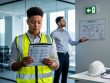 Two colleagues in a modern workplace wearing safety vests review workplace safety materials, with personal protective equipment visible on a table in the background.